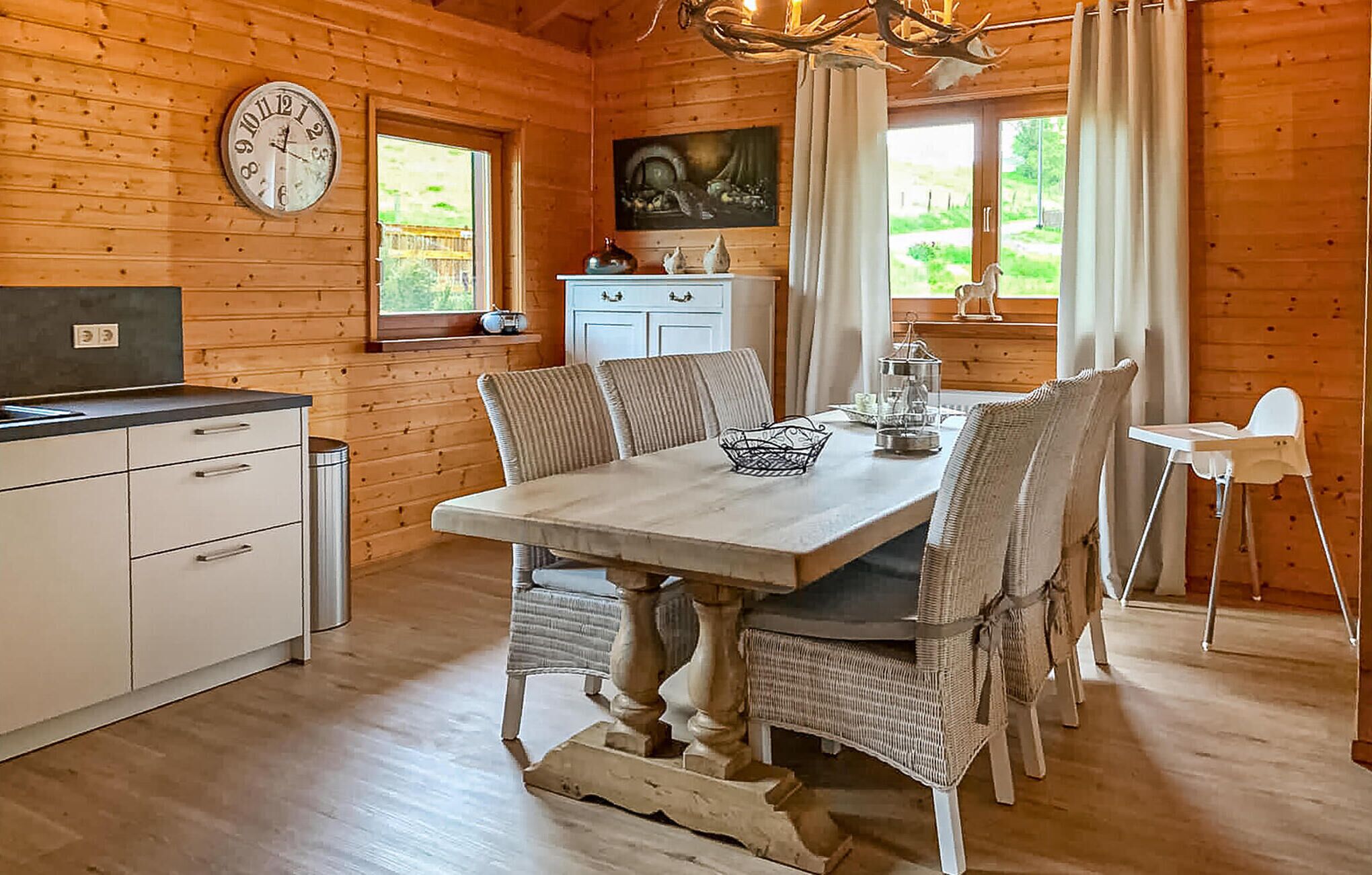 Interior de una cabaña de madera con una mesa de comedor, sillas, un reloj y una zona de cocina.