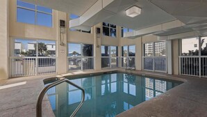 An indoor swimming pool with clear glass walls, a metal pool ladder, and a view of palm trees and buildings outside. - Dog friendly beachfront condo with in/out pools (Gulf Shores)