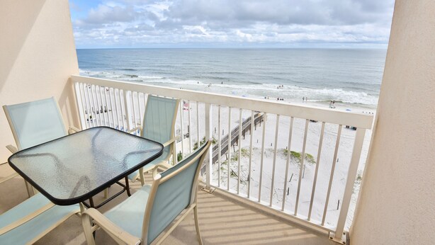 A balcony with a glass table and chairs, overlooking a beach with a pier and people. - Dog friendly beachfront condo with in/out pools (Gulf Shores)