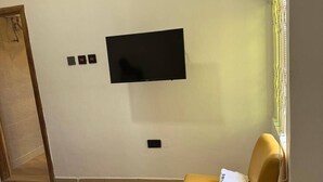 A hotel room with a bed, a yellow armchair, a TV mounted on the wall, and a window with blinds. - Cosy Rooms Away in a Stunning House (Koforidua)