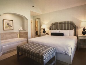 Een hotelkamer met een groot bed, een klein bankje, een nachtkastje met een lamp, een spiegel en een lijst met een afbeelding aan de muur.