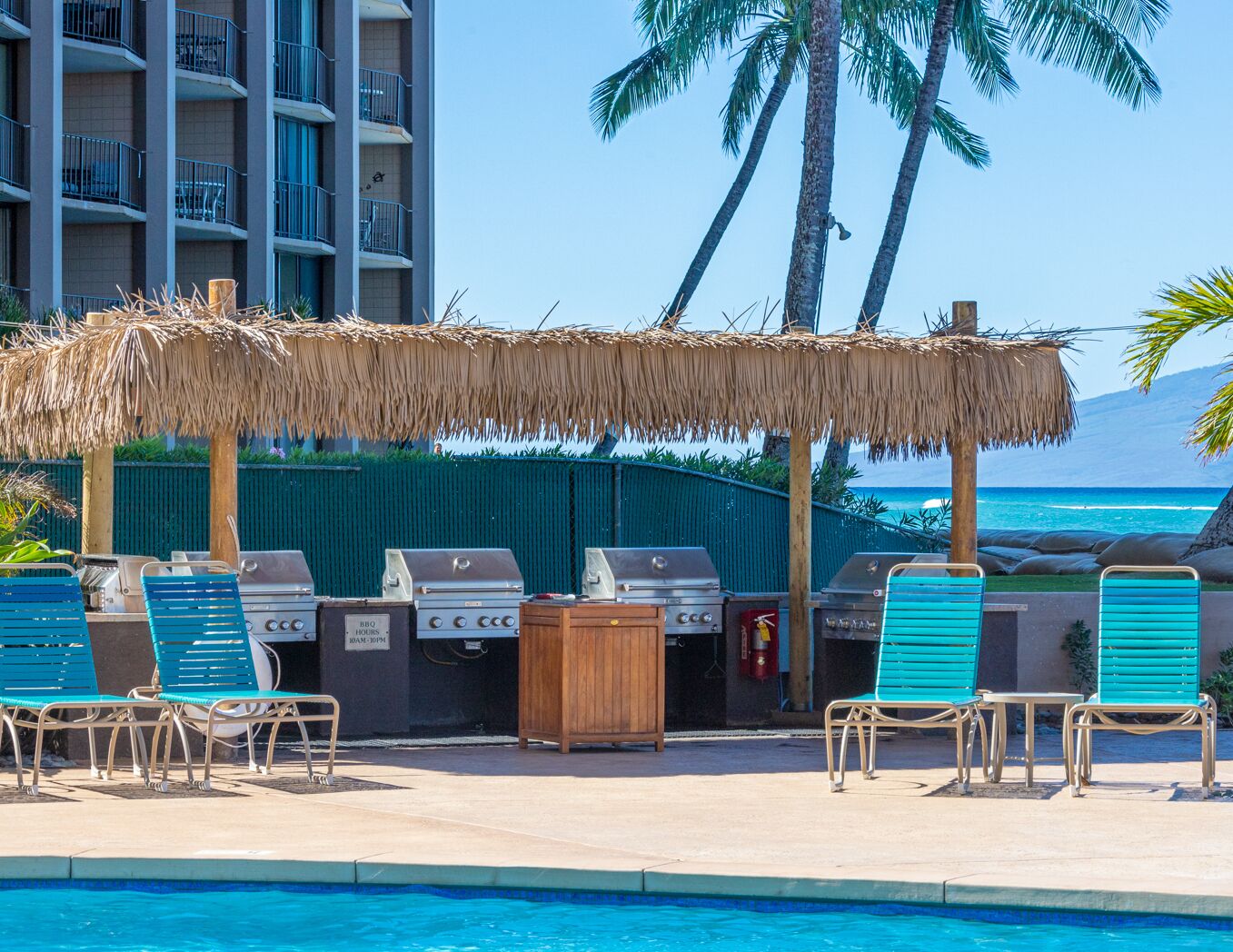 A poolside area with a barbecue grill, lounge chairs, and a thatched umbrella.