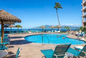 A pool area with lounge chairs and umbrellas, a clear view of the ocean, and a mountain in the background.