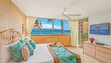 Um quarto com cama, ventilador de teto, vista para o oceano, televisão na parede e cômoda de madeira.