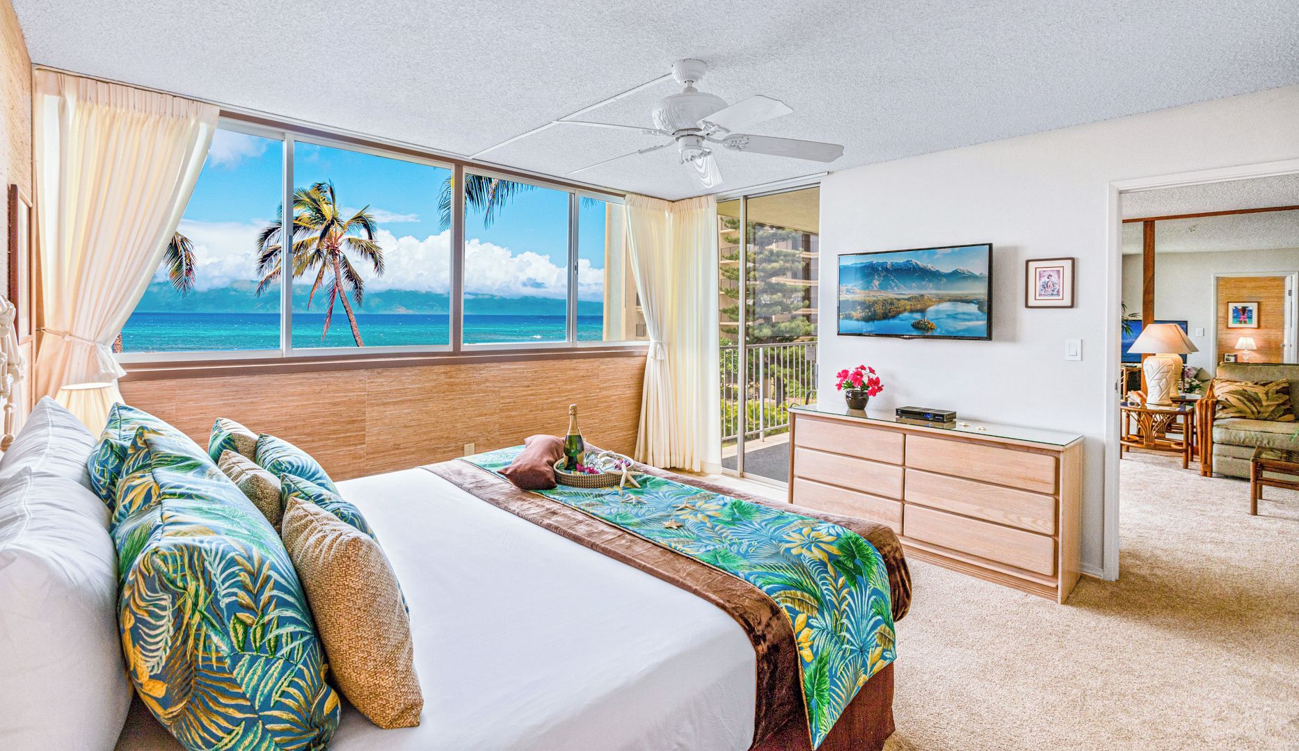 A bedroom with a large window overlooking a beach, a bed with tropical-themed pillows, a ceiling fan, and a small desk with a lamp.