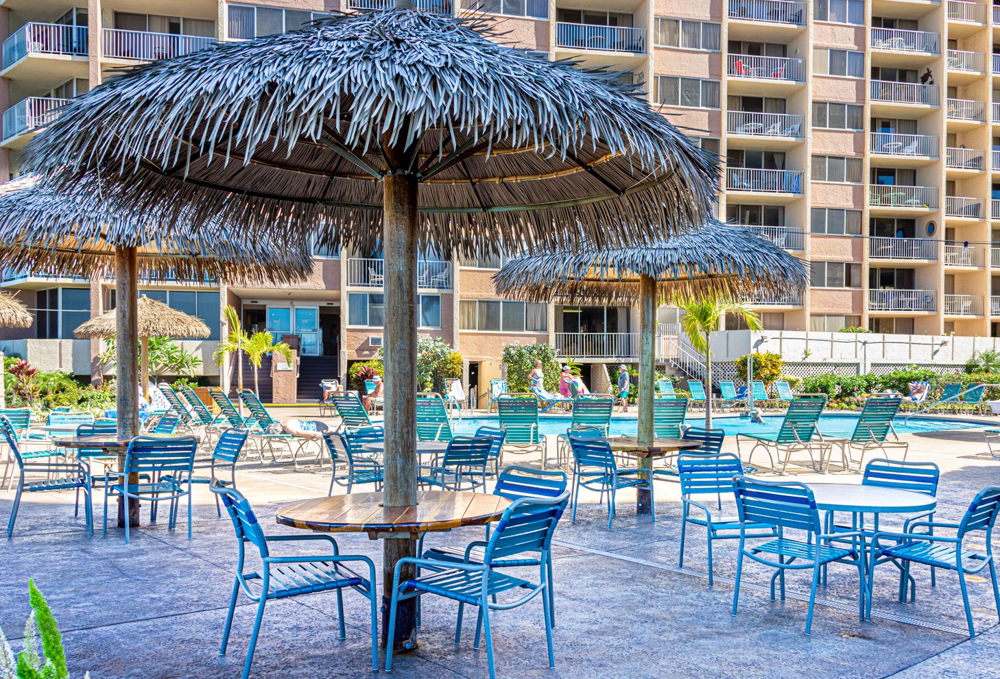 A poolside area with thatched umbrellas, lounge chairs, and a multi-story building in the background.