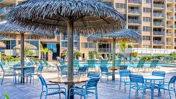 A poolside area with thatched umbrellas, lounge chairs, and a multi-story building in the background.