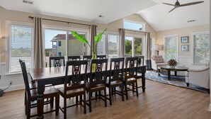 Un vaste espace dédié à la salle à manger, avec une grande table et des chaises en bois, un ventilateur de plafond et de grandes fenêtres équipées de stores.