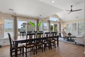 A spacious dining area with a large wooden table and chairs, a ceiling fan, and large windows with blinds.