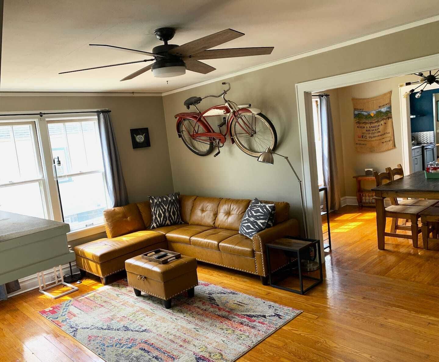 A living room with a brown leather sofa, a small ottoman, a colorful rug, a ceiling fan, and a bicycle mounted on the wall.