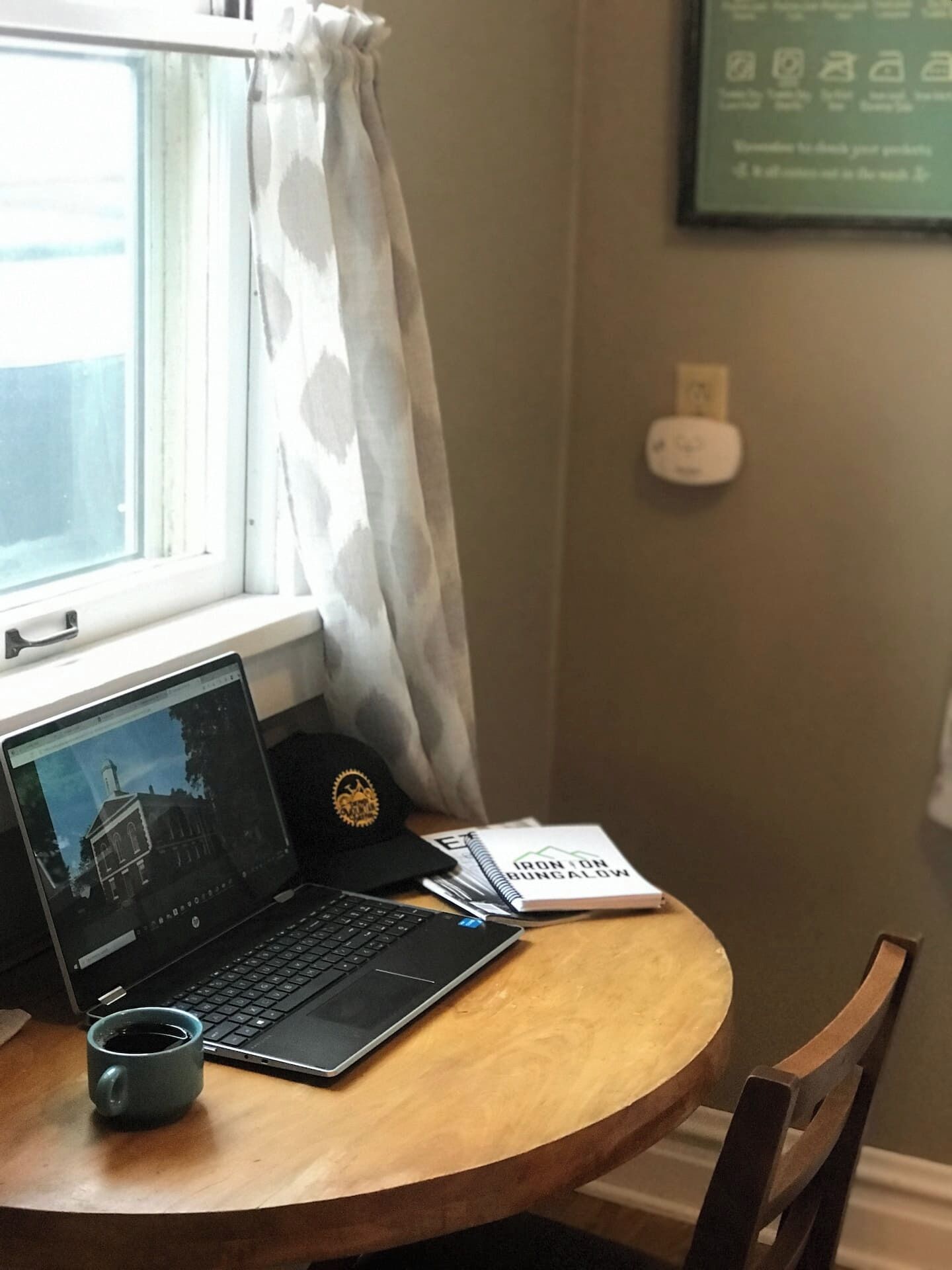 A laptop on a wooden table near a window with a curtain, a notebook, a smartphone, and a mug.