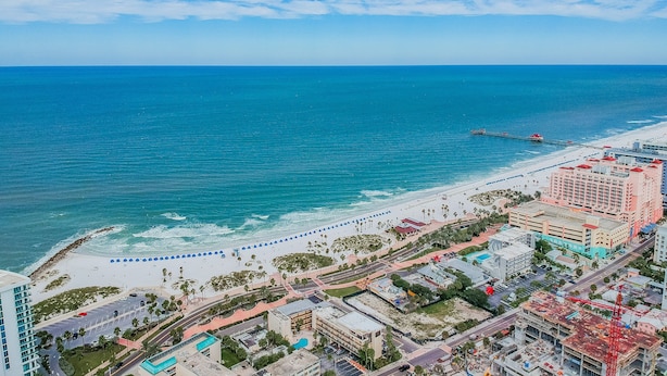 Paisagem costeira com praia, um cais e um hotel com vários andares e varandas.