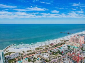 Un paisaje urbano costero con playa, muelle y un hotel de varios pisos con balcones.