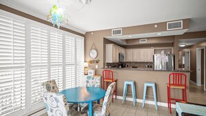 A modern kitchen with white cabinets, a stainless steel refrigerator, and a microwave. There is a dining area with a blue table and patterned chairs.