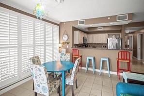A modern kitchen with white cabinets, a stainless steel refrigerator, and a microwave. There is a dining area with a blue table and patterned chairs.