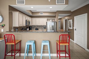 A kitchen with white cabinets, a stainless steel refrigerator, a microwave, and a granite countertop. There are two red chairs and two blue stools.