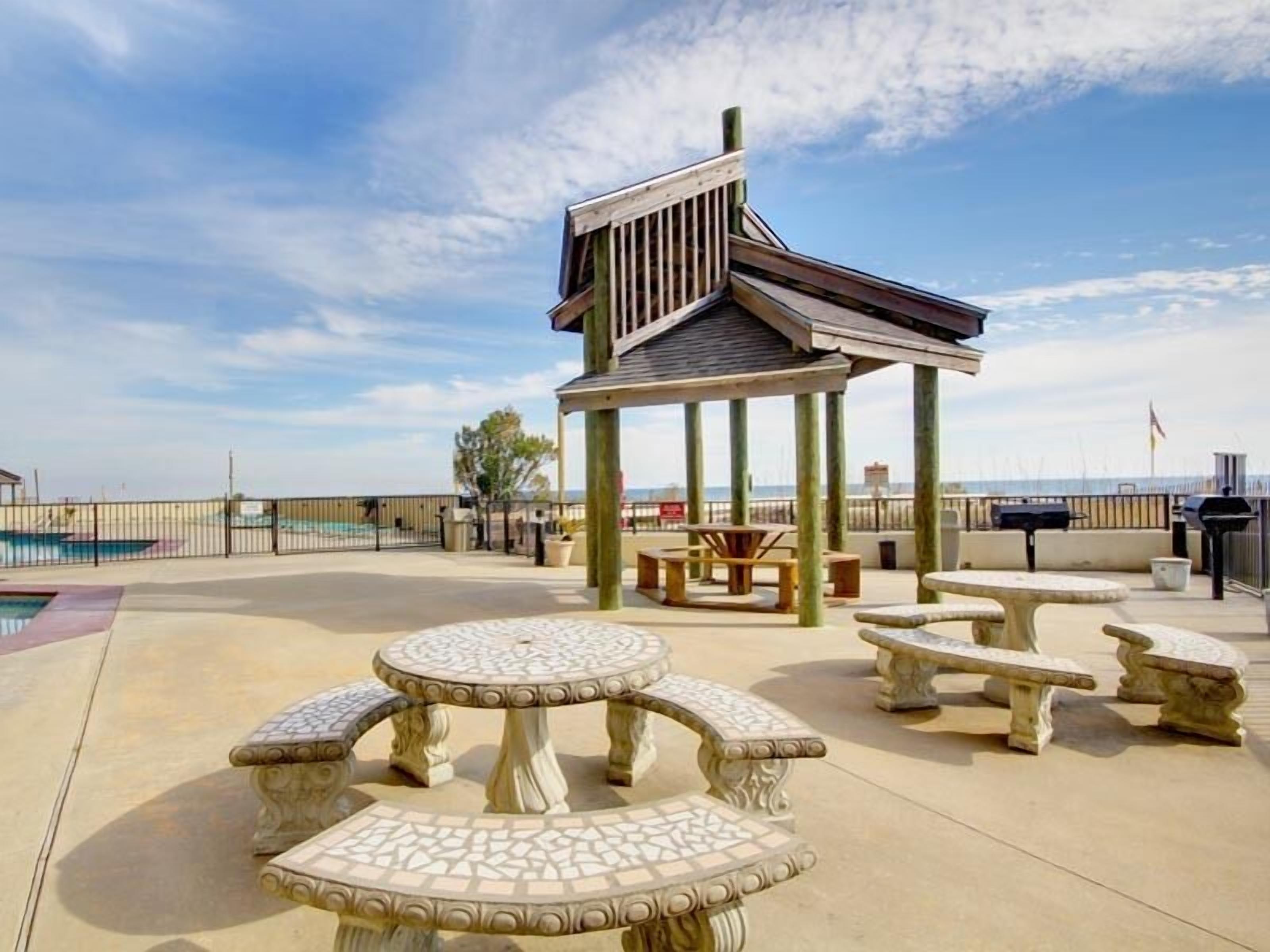 An outdoor patio area with a gazebo, multiple stone tables, and a pool in the background.