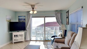 A living room with a TV, sofa, coffee table, and a balcony with outdoor furniture. - Regency Towers #508 West | Gulf Views, Pool! (Pensacola Beach)