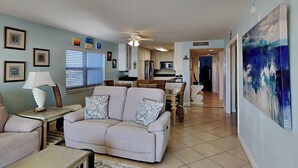 A living room with a sofa, a coffee table, and a lamp. There is a kitchen in the background with cabinets and a microwave. - Regency Towers #508 West | Gulf Views, Pool! (Pensacola Beach)