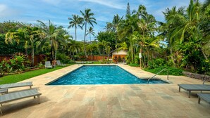 Piscina retangular com deck revestido, rodeada por palmeiras e vegetação exuberante.
