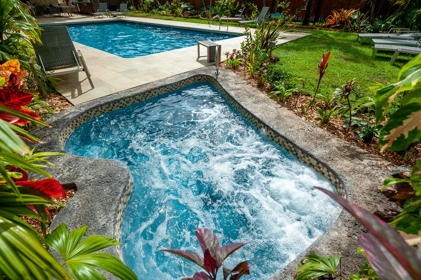 Uma banheira de hidromassagem retangular com borda em azulejo, rodeada por vegetação exuberante e uma piscina ao fundo.