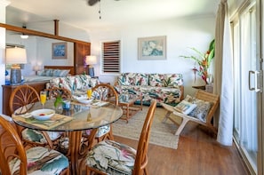 A dining area with a round glass table, wooden chairs, and a floral-patterned sofa. - Quaint Island Charm Right On the Beach! Secluded & Laidback Seaside Retreat (Hanalei)