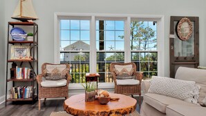 A living room with two wicker chairs, a round coffee table, a bookshelf, and a large window with a view of trees. - Designer Home, New Gulf View Home with Comm. Pool ~ Bethany Cottage (Indian Pass)