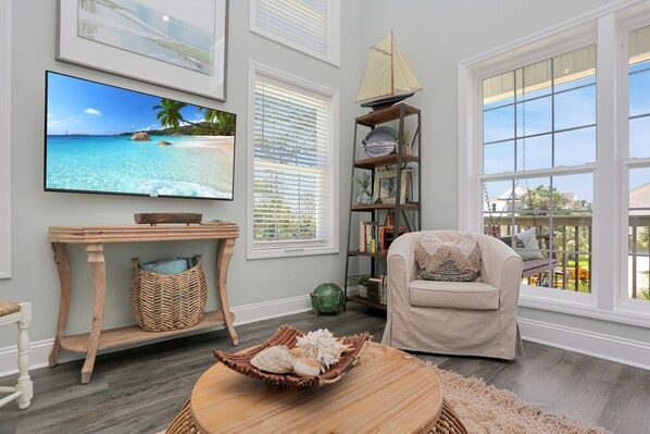 A living room with a large TV, a bookshelf, a wicker basket, and a wicker chair. - Designer Home, New Gulf View Home with Comm. Pool ~ Bethany Cottage (Indian Pass)