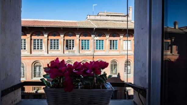 Un balcó amb una cistella de vimet plena de flors, vista d'un edifici de maó vermell amb finestres d'arc i un cel blau clar.