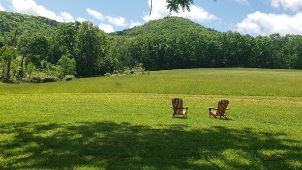 Un campo cubierto de césped con dos sillas Adirondack mirando una colina boscosa bajo un cielo despejado.