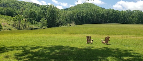 A grassy field with two Adirondack chairs facing a forested hill under a clear sky.