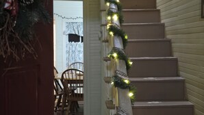 Una escalera adornada con luces y ramas de pino, que conduce a un comedor con una mesa y sillas de madera.