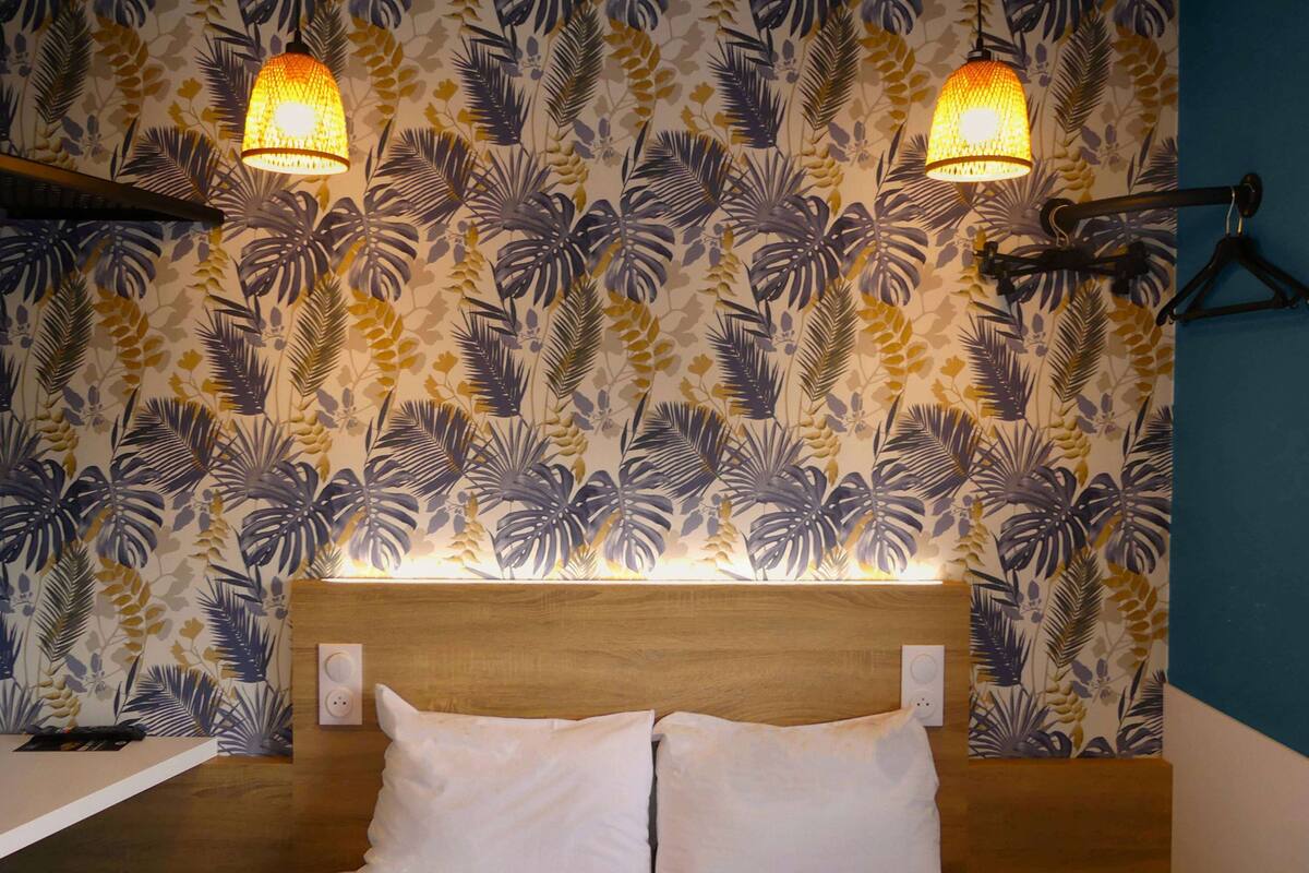 A hotel room with a bed, two wall-mounted lamps, and a tropical leaf-patterned wallpaper.