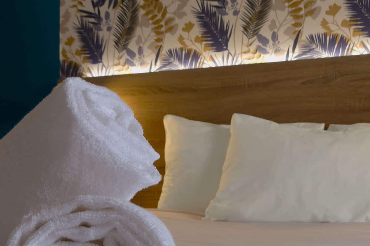 A neatly made bed with white linens and pillows, a rolled white towel, and a wall with a tropical leaf pattern.