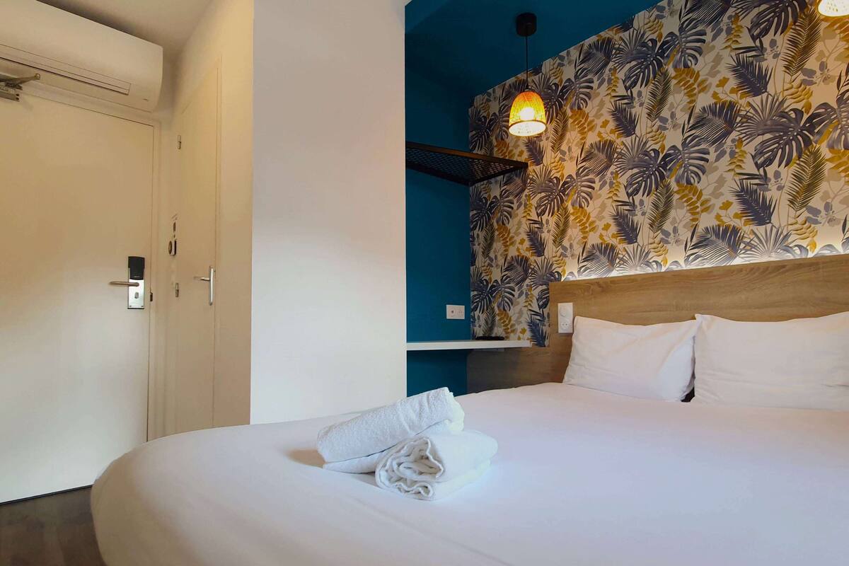 A hotel room with a neatly made bed, a wall with tropical leaf wallpaper, and a ceiling light fixture.