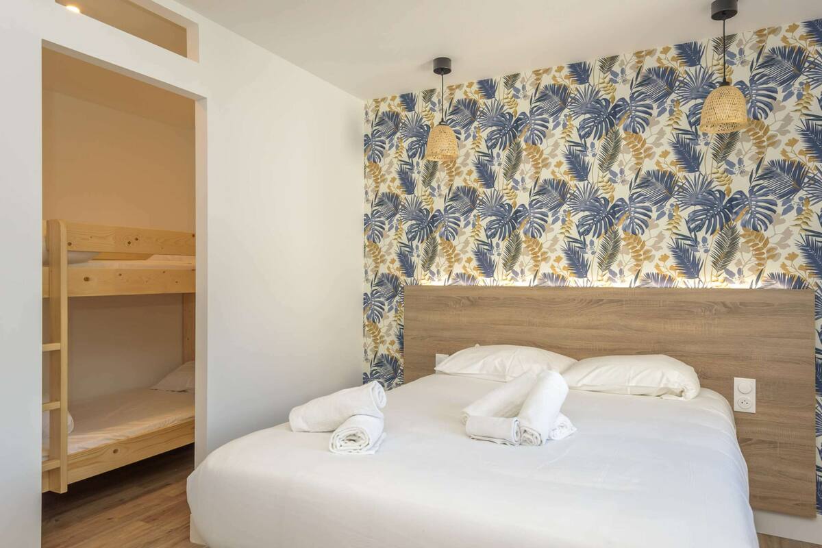 A bedroom with a bunk bed, a large bed with neatly folded towels, and a wall with a tropical leaf pattern.
