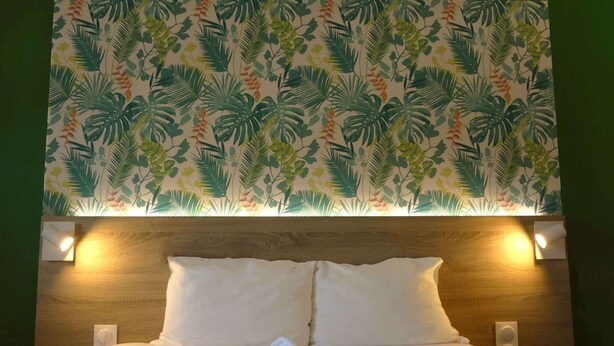 A hotel room with a bed, two pillows, a folded towel, and a wall with a tropical leaf pattern. - Kyriad ECO - Toulon Ouest La Seyne sur Mer (La Seyne-sur-Mer)