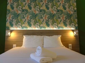 A hotel room with a bed, two pillows, a folded towel, and a wall with a tropical leaf pattern. - Kyriad ECO - Toulon Ouest La Seyne sur Mer (La Seyne-sur-Mer)