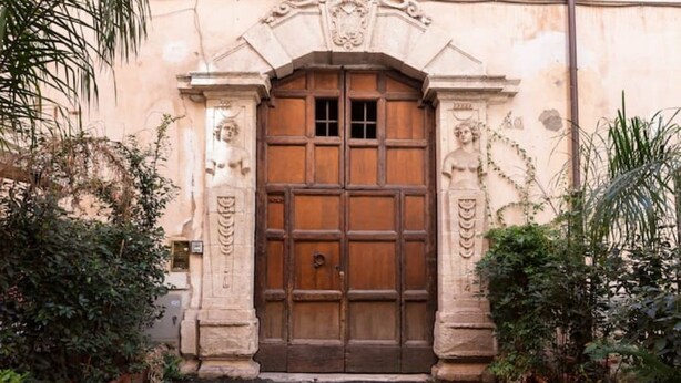 Una gran puerta de madera con intrincados grabados, flanqueada por pilares de piedra con detalles decorativos, ubicada en un jardín con plantas en macetas.