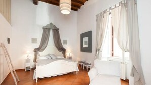 Un dormitorio con cama, mesitas de noche, una silla, una ventana con cortinas y un cuadro en la pared.