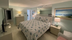 Ein Schlafzimmer mit einem Bett, Nachttischen, einem Fernseher, einem Gemälde und einer Tür zu einem anderen Raum.