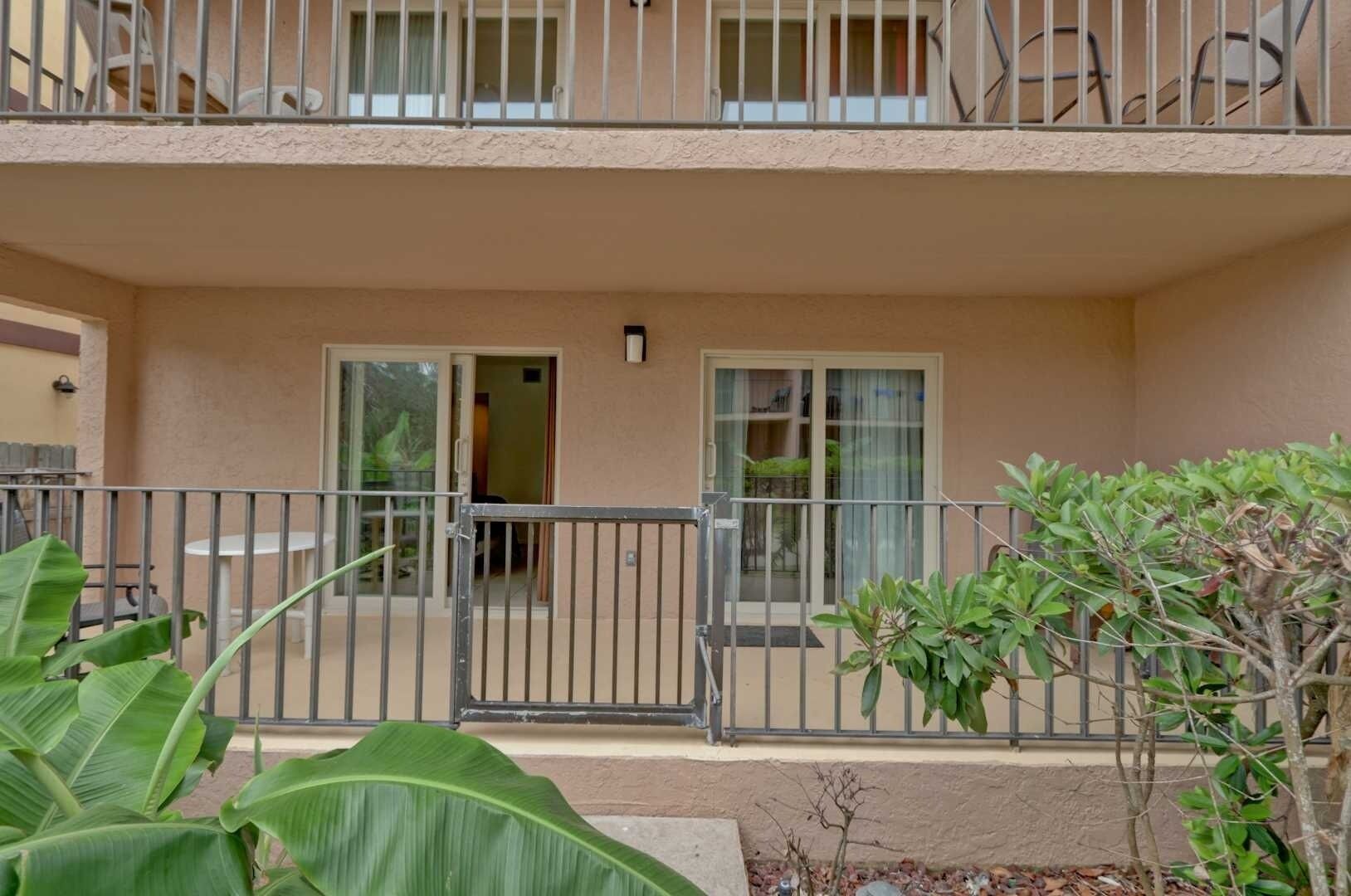 A two-story residential building with a balcony, a sliding glass door entrance, and a small garden area with a metal fence.