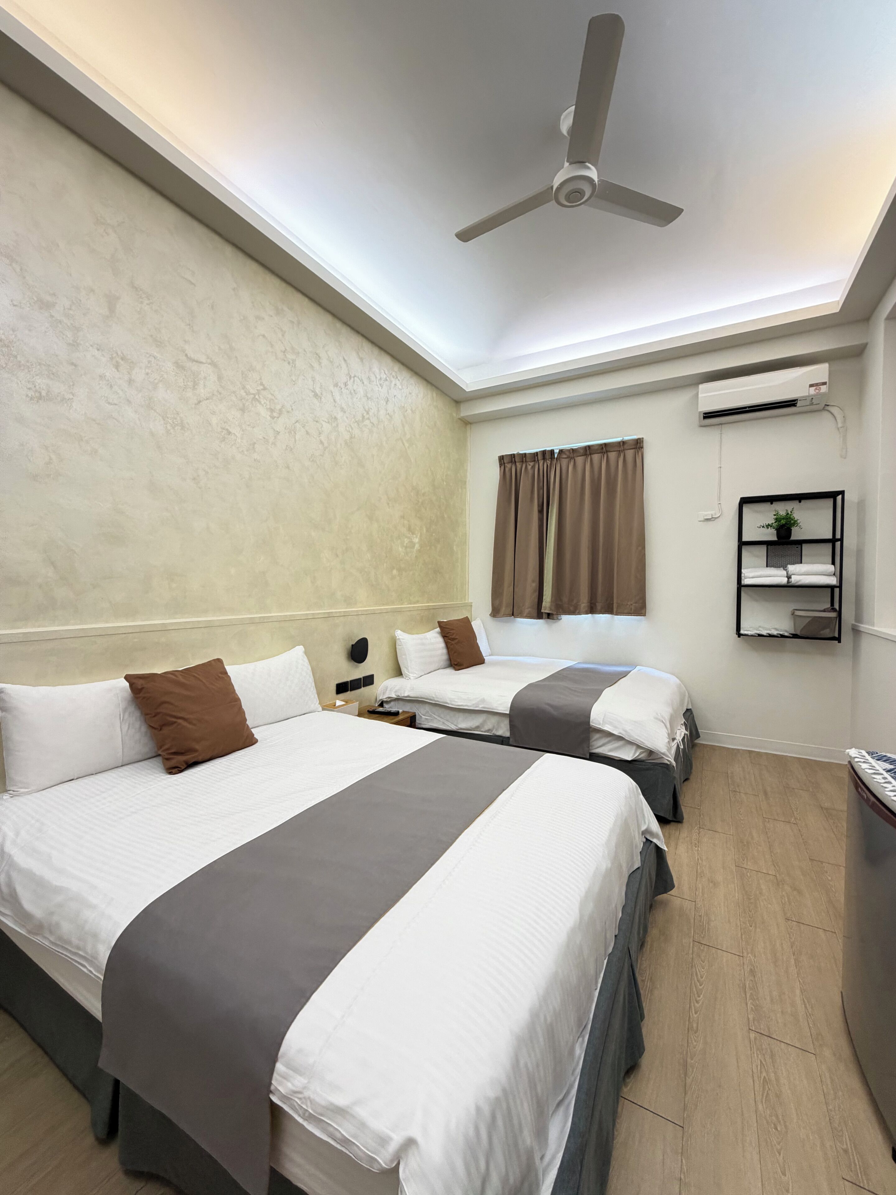 A hotel room with two beds, a ceiling fan, an air conditioning unit, a shelf with a plant, and a window with curtains.