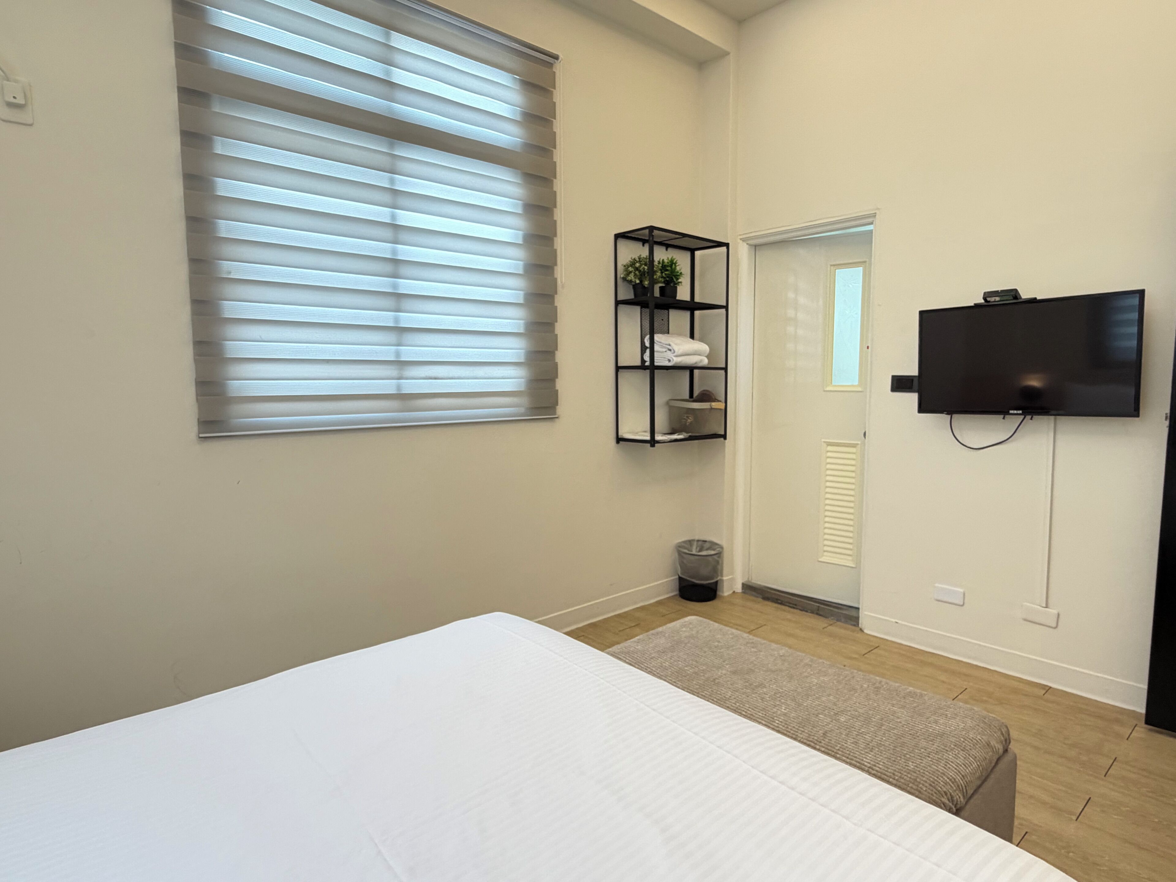 A bedroom with a bed, a TV mounted on the wall, a window with blinds, a shelf with towels, and a trash bin.
