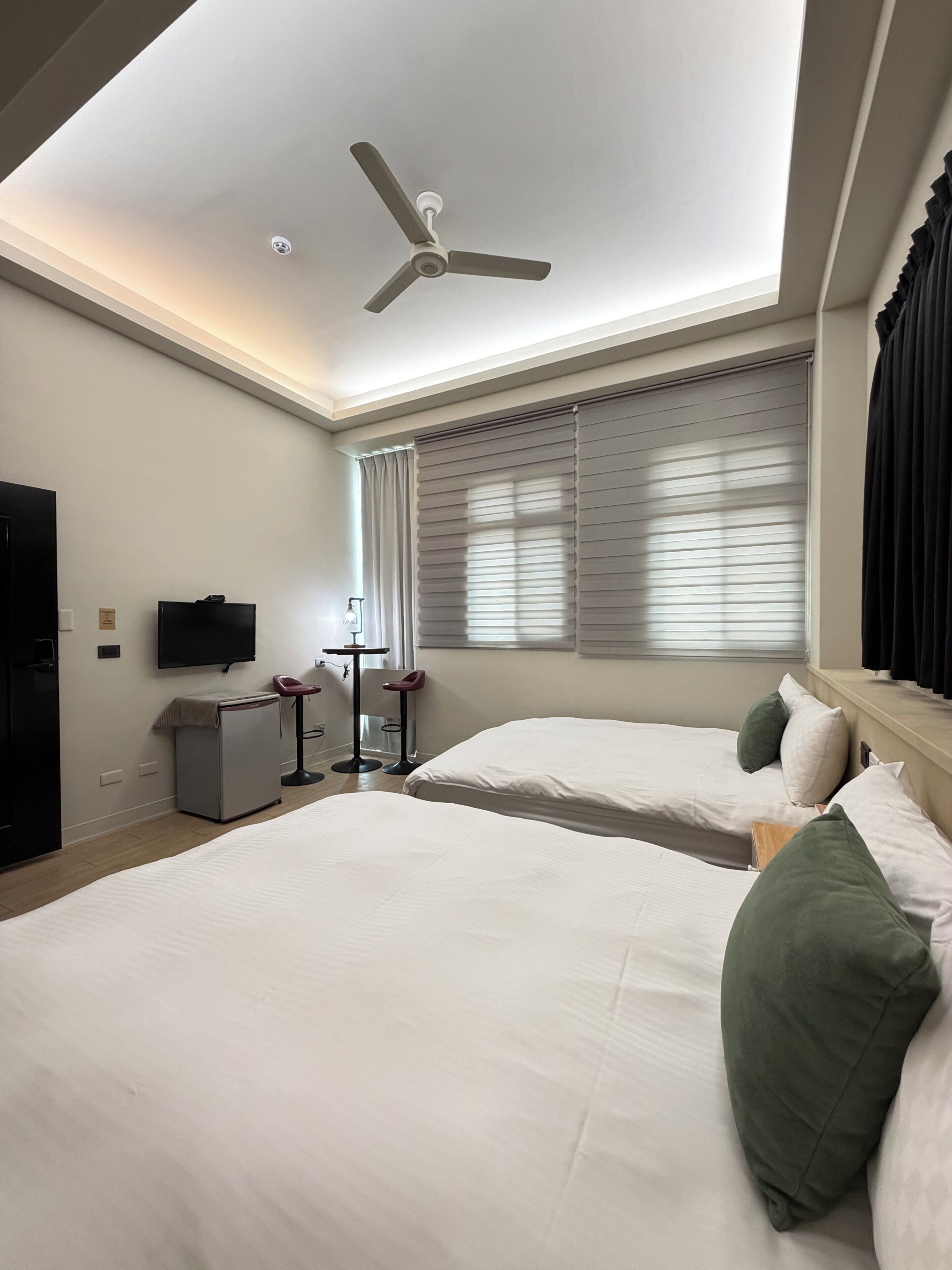 A hotel room with two beds, a ceiling fan, a TV, a minibar, a small table with a chair, and a window with blinds.