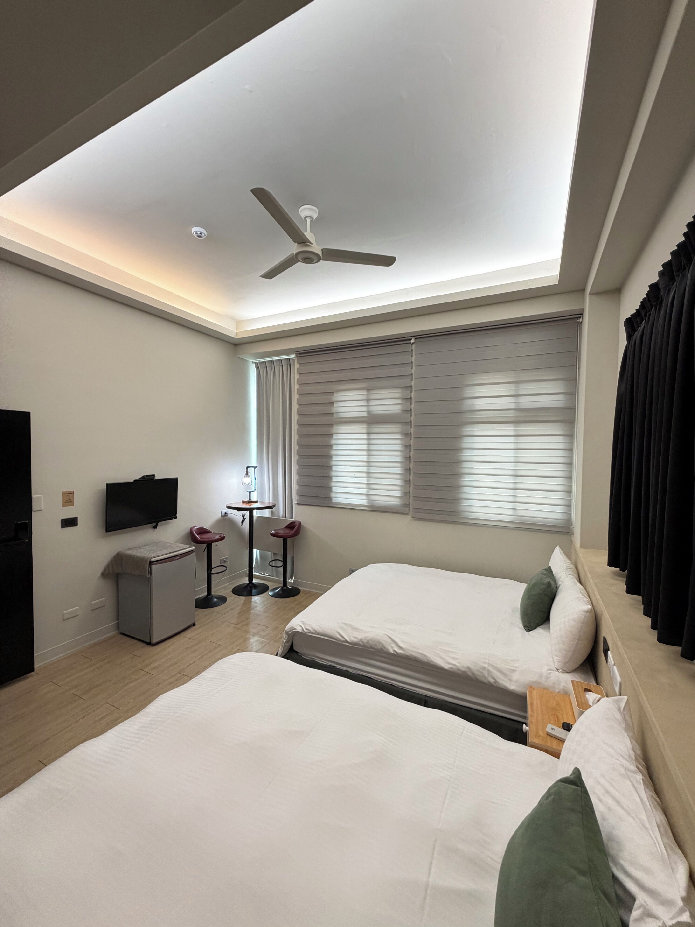 Une chambre d’hôtel avec deux lits, un ventilateur de plafond, une télévision, une petite table avec une chaise et une fenêtre avec des stores.