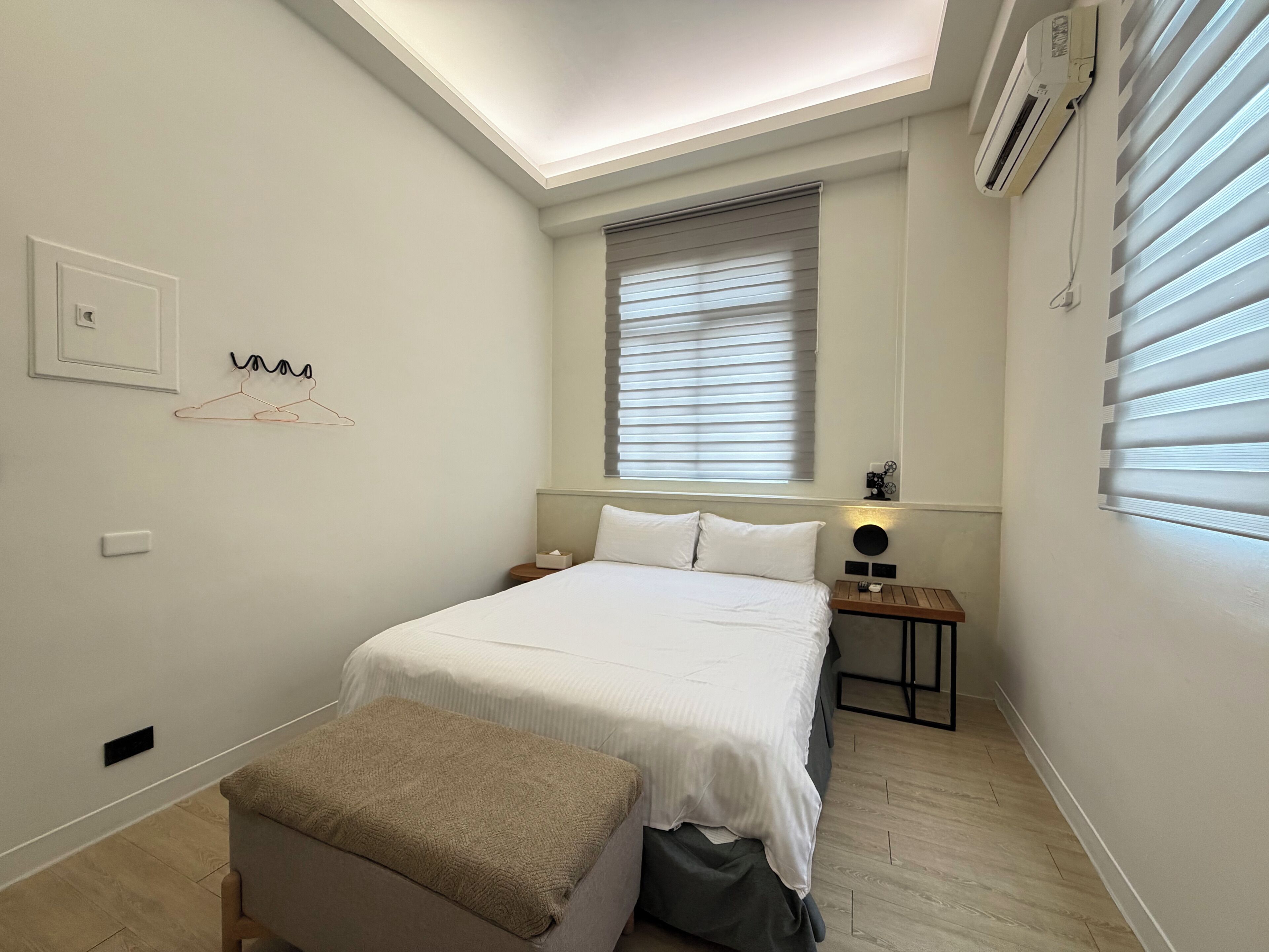 A bedroom with a bed, a bedside table, a bench, a window with blinds, an air conditioning unit, and a wall-mounted light fixture.