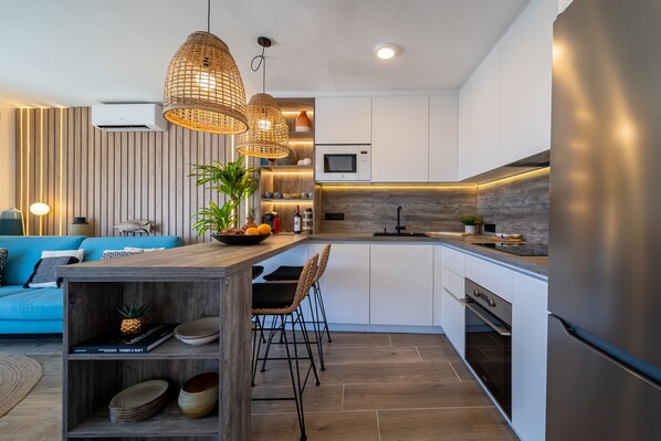A modern kitchen with a central island, white cabinetry, and stainless steel appliances. There is a dining area with a blue sofa and a wooden coffee table. - Casa Galana | Stylish apartment on the seafront (Tías)