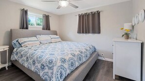 Una camera da letto con un letto, una ventola a soffitto, una finestra con tende, un comodino e un mobile con una pianta.