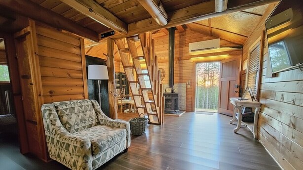 Interno di una baita in legno con una poltrona a fantasia, pavimento in legno, un caminetto e una scala che conduce a una zona mansardata.